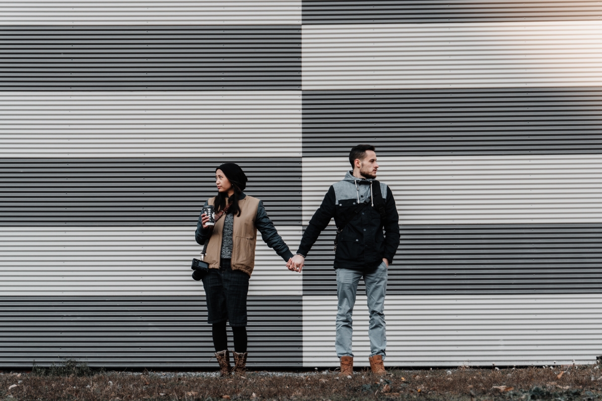 man and woman holding hands in front of grey and white panelled wall. they are looking on opposite directions.