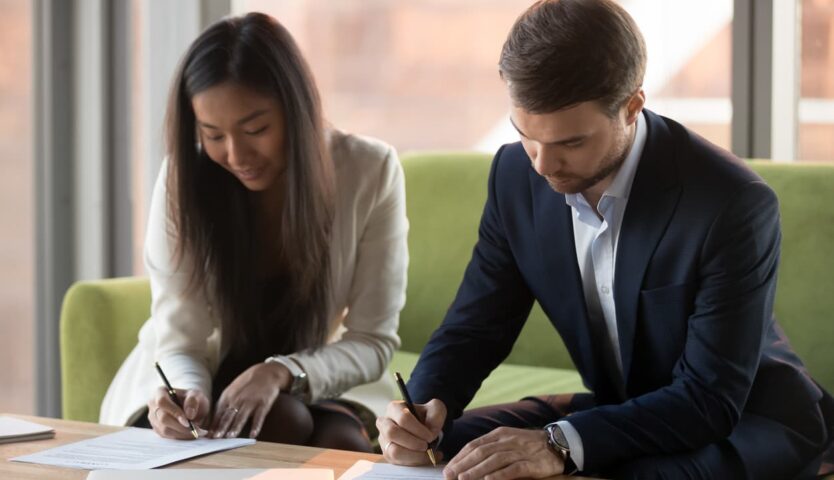 Two business professionals seated together in an office, reviewing and signing paperwork on a table, representing contract agreements, business partnerships, or financial planning.