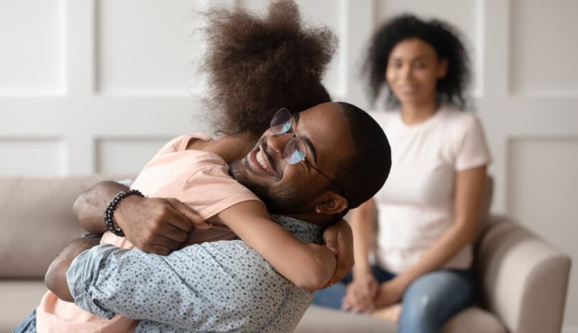 A smiling father embracing a young child in a living room while another parent watches from a sofa in the background, capturing a moment of family affection and connection.