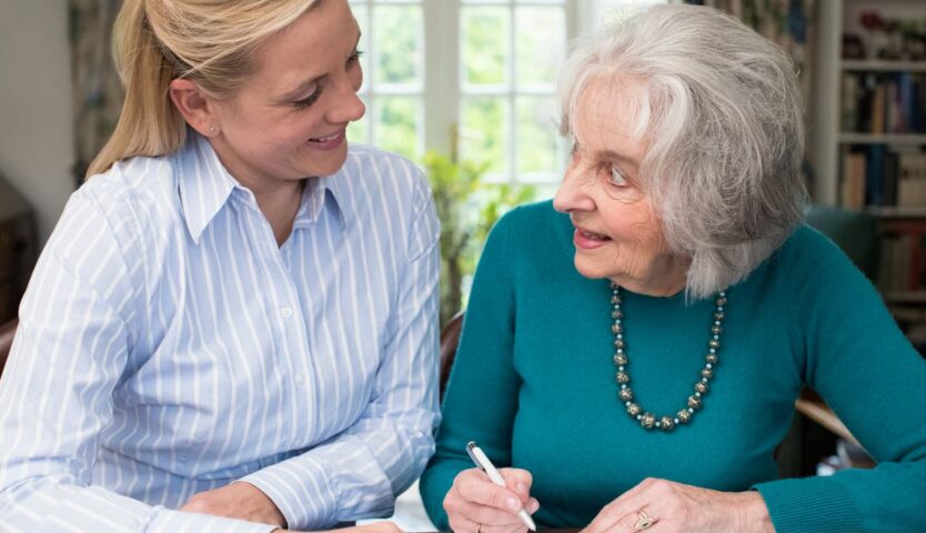 A caregiver sitting beside an elderly woman as they review and complete documents together at home, representing senior care, support services, and assistance with personal affairs.