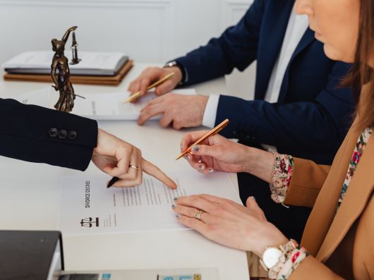 A lawyer points to a section on a divorce decree document while two clients review and sign paperwork at a desk, with legal files and a Lady Justice statue nearby.