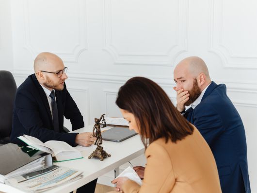 A lawyer in a suit meeting with two clients at a desk, discussing legal documents in an office setting with a small Lady Justice statue on the table.