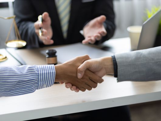 Two clients shake hands in agreement during a meeting with a divorce lawyer, with legal scales and a gavel on the desk in the background.